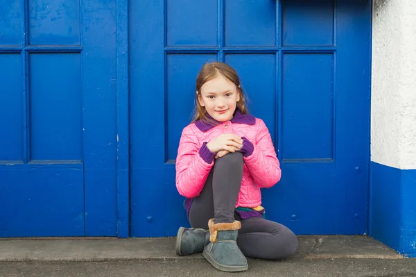 Fashion portrait of a cute little girl, sitting on the floor next to bright blue door, wearing pink coat and blue boots — Stock Photo, Image