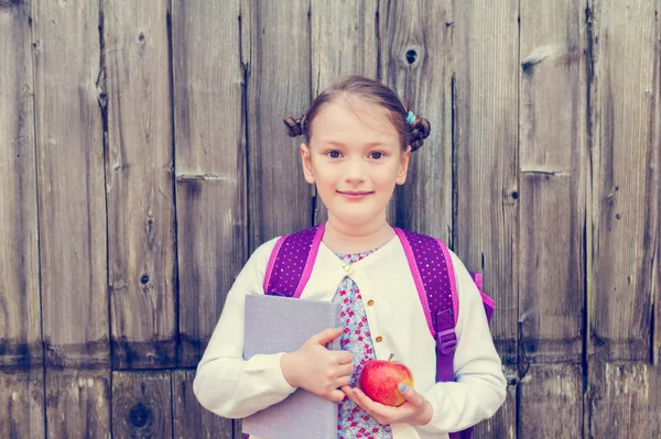 Cute little girl with backpack, holding a book and red apple, standing against wooden background, back to school concept, toned image, vintage effect — Stock Photo, Image