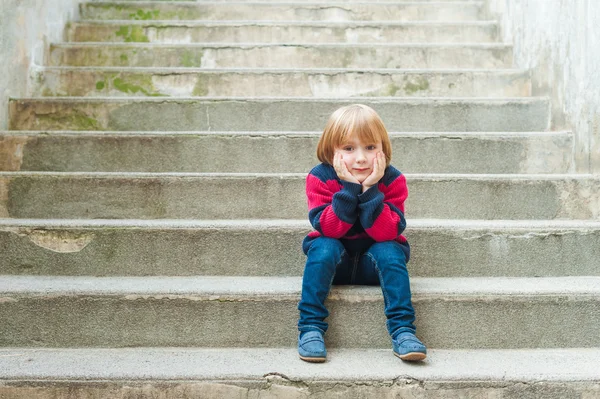 Adorable little boy with blond hair sitting on steps in a city on a nice sunny day, wearing warm pullover, jeans and blue moccasins — Stock Photo, Image