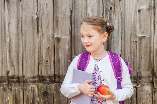 Retrato ao ar livre de uma menina bonita de 7 anos de idade, usando mochila, segurando um livro e maçã vermelha, de pé na frente da parede de madeira velha, de volta ao conceito da escola — Fotografia de Stock