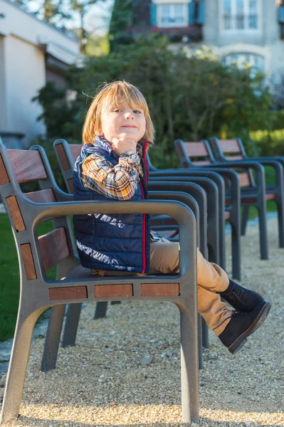 Zabawny małego chłopca spoczywa na ławce w parku, na sobie niebieską kamizelkę i buty — Zdjęcie stockowe
