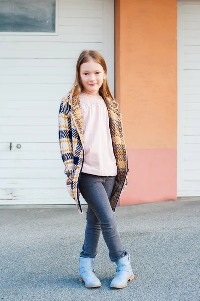 Outdoor portrait of a cute little girl, wearing plaid cardigan, grey jeans and blue boots, standing next to white wooden wall — Stock Photo, Image