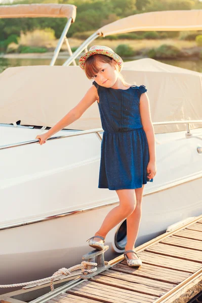 Sunset portrait of a cute little girl, standing by the lake next to white yacht, wearing blue, dress and colorful hat — Stock Photo, Image