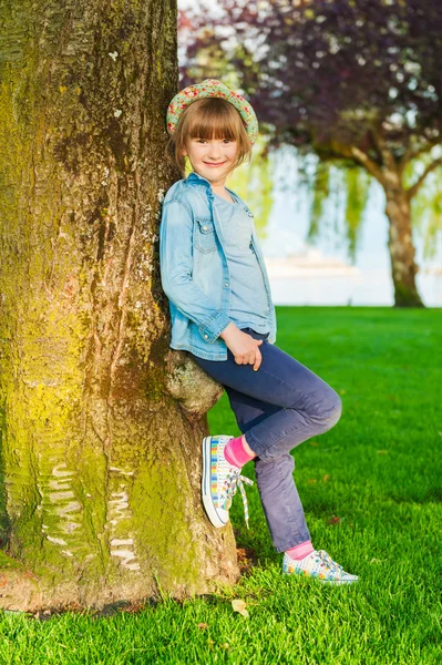 Summer portrait of a cute little girl of 6 years old, wearing blue clothes, colorful hat and shoes, standing next to tree in the park — Stock Photo, Image