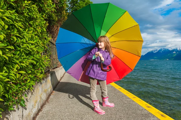 Outdoor portrait of a cute little girl with big colorful umbrella, wearing purple rain coat, boots, standing next to lake — Stock Photo, Image