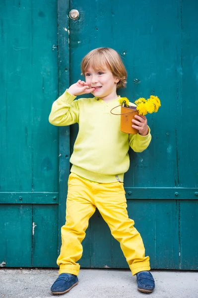 Außenporträt eines niedlichen kleinen Jungen in gelbem Pullover und Hose, der an der grünen Holztür steht und einen Eimer mit Löwenzahn in der Hand hält. — Stockfoto