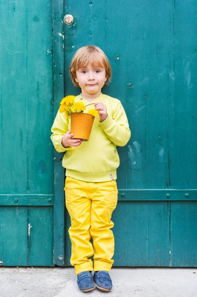 かわいい小さな男の子が、黄色のセーターとズボンを着て、緑の木製のドアのそばに立って、タンポポの花とバケツを持っての屋外のポートレート — ストック写真