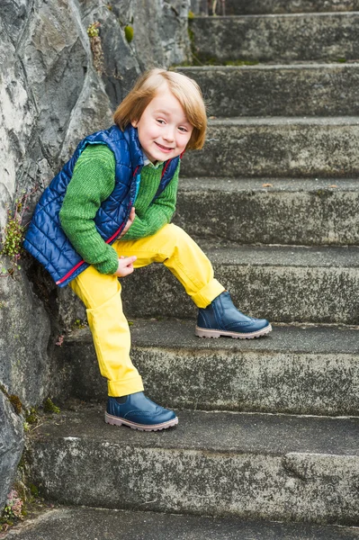 Outdoor portrait of a cute little blond boy wearing colorful clothes, yellow jeans, green pullover, blue waistcoat and boots, standing on stairs in a city — Stock Photo, Image