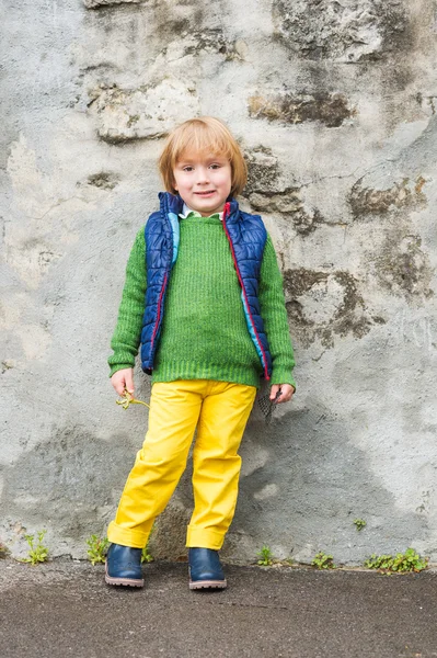 Outdoor portrait of a cute little blond boy wearing colorful clothes, yellow jeans, green pullover, blue waistcoat and boots, standing next to stone wall in a city — Stock Photo, Image