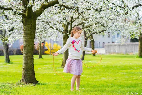 Menina bonita brincando em um jardim de primavera em um dia ensolarado agradável, pulando com corda, vestindo sweatdhirt com um coração e saia rosa — Fotografia de Stock