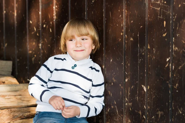 Ładny mały kaukaski blond chłopiec opierał się na starej drewnianej ścianie, noszenie biały sweter — Zdjęcie stockowe