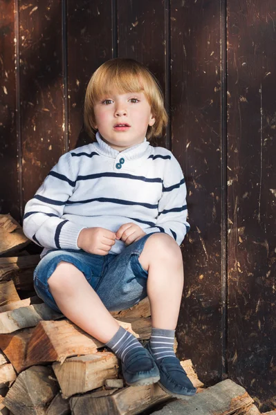 Schattige kleine blonde jongen zittend op gesneden hout en leunend op een oude houten muur, dragen witte pullover, denim shorts en blauwe mocassins — Stockfoto