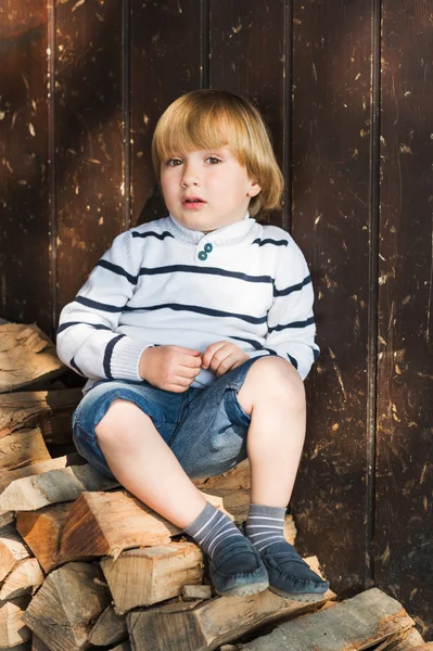 Cute little blond boy sitting on cut wood and leaning on an old wooden wall, wearing white pullover, denim shorts and blue moccasins — Stock Photo, Image