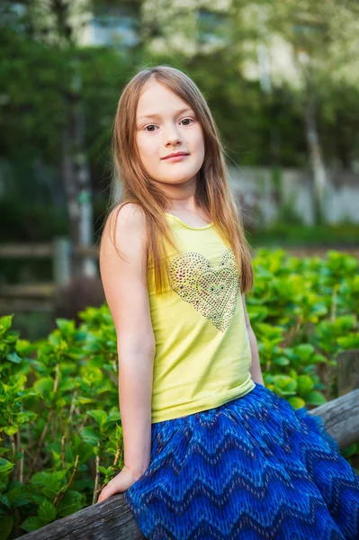 Summer portrait of a pretty little girl, sitting on a fence at sunset, wearing green t-shirt and blue skirt — Stock Photo, Image