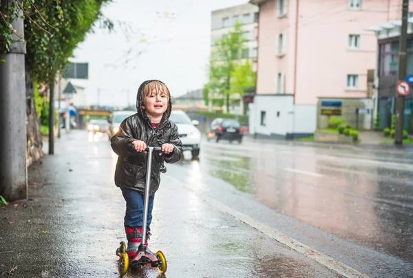 Cute little boy in a very wet clothes on the scooter next to road under the rain — Stock Photo, Image