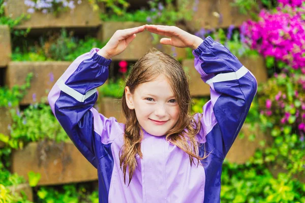 Außenporträt eines niedlichen kleinen Mädchens im Regen, das lila Regenmantel trägt und die Hände über dem Kopf hält — Stockfoto