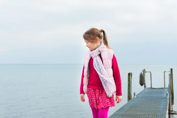 Милая маленькая девочка играет у озера, ходит по пирсу, в розовом жилете — стоковое фото