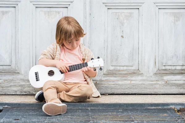 Pequeño niño feliz toca su guitarra o ukelele, sentado junto a la puerta de madera al aire libre — Foto de Stock