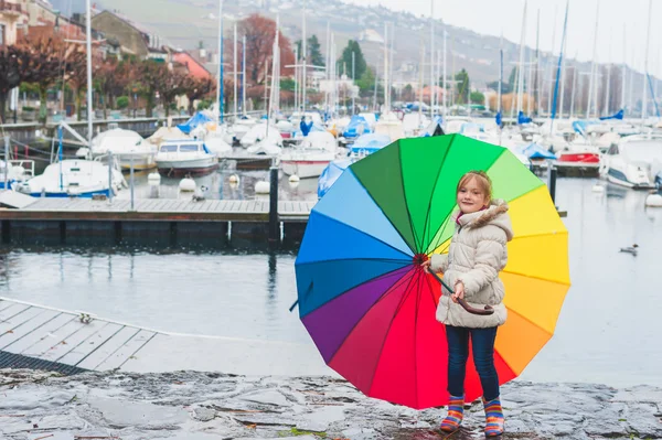 Outdoor portrait of a cute little girl of 6 years old with big colorful umbrella, playing by the lake on a cold rainy day, wearing warm white coat and rainbow boots — Stock Photo, Image