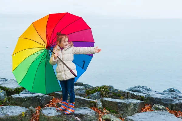 Outdoor portrait of a cute little girl of 6 years old with big colorful umbrella, playing by the lake on a cold rainy day, wearing warm white coat and rainbow boots — Stock Photo, Image