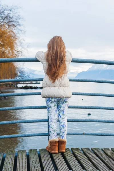 Niedliches kleines Mädchen, das auf einer Brücke steht, auf den schönen See blickt, einen warmen beigen Mantel trägt, Rückansicht — Stockfoto