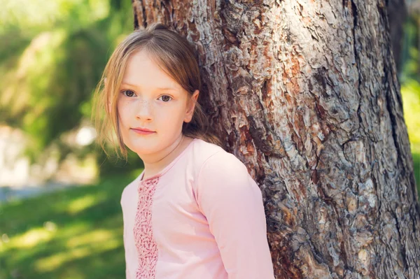 Outdoor portrait of a cute little girl of 7 years old, resting in a park, leaning on a tree — Stock Photo, Image