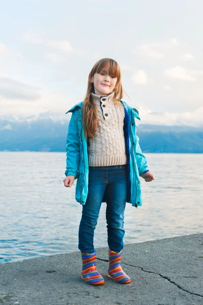 Retrato al aire libre de una linda niña descansando junto a los lkae, con abrigo azul y botas de lluvia coloridas — Foto de Stock