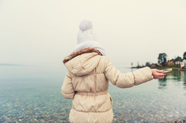 Little girl playing by the lake, throwing stones into the water on a nice cold day, wearing warm white hat, scarf and beige coat, back view, toned image