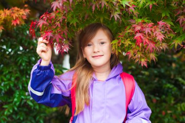 Autumn portrait of adorable little girl wearing purple rain coat and red backpack