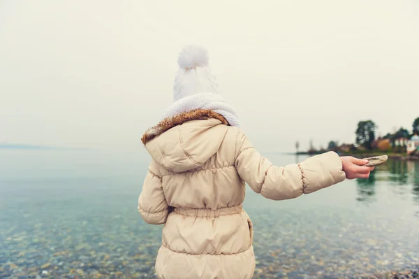 Menina brincando junto ao lago, jogando pedras na água em um dia frio agradável, vestindo chapéu branco quente, cachecol e casaco bege, visão traseira, imagem tonificada — Fotografia de Stock