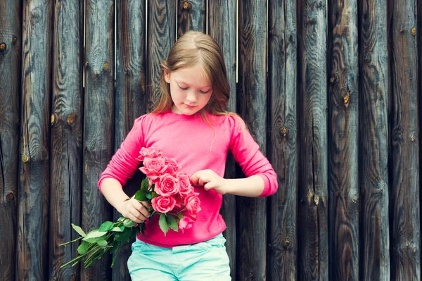 Outdoor portrait of a cute little girl against wooden wall, wearing bright pink pullover, holding bouquet of roses — ストック写真