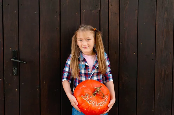 Outdoor portrait of a cute little girl holding big pumpkin with french sign Bienvenue (welcome)