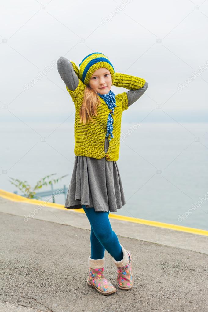 Cute little girl having fun outdoors on a cold day, dancing next to beautiful lake, wearing green woolen pullover, hat and warm boots