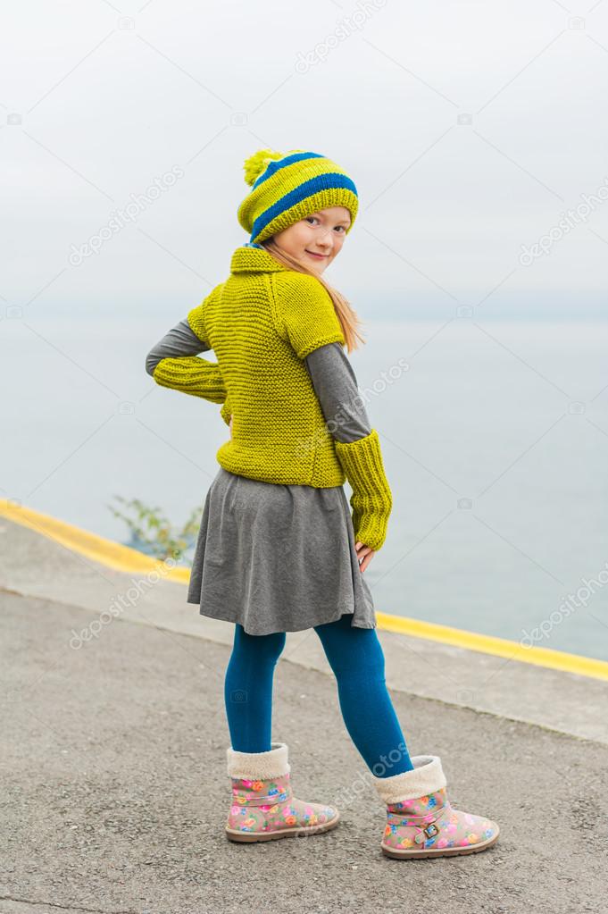 Cute little girl having fun outdoors on a cold day, dancing next to beautiful lake, wearing green woolen pullover, hat and warm boots