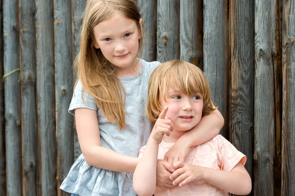 Outdoor portrait of two cute kids, big sister and her little brother against wooden wall — Stock Photo, Image