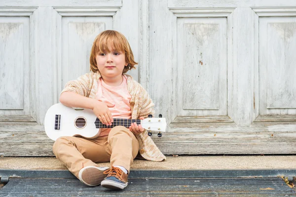 Szczęśliwy chłopiec gra jego gitara lub ukulele, siedząc przez drewniane drzwi na zewnątrz — Zdjęcie stockowe