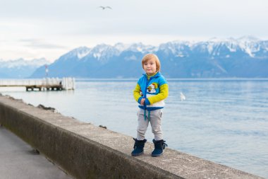 Winter portrait of a cute little boy, wearing funny warm knitted jacket with snowman, standing next to lake against mountains