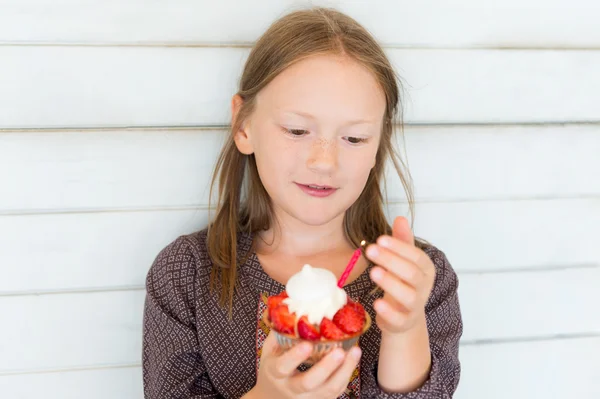 Close up portrait of a cute little girl of 7-8 years old, standing against white wooden background, holding small strawberry cake with one candle — Stock Photo, Image