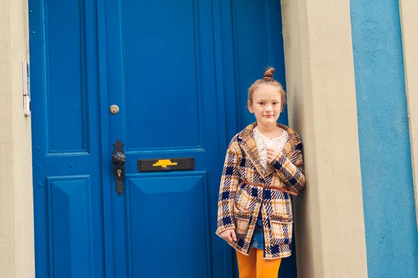 Ritratto di moda di una bambina carina, con indosso un caldo cardigan a maglia, in piedi accanto alla porta blu brillante — Foto Stock