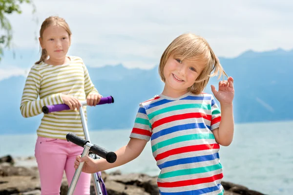 Two cute kids playing by the lake, holding their scooters — Stock Photo, Image