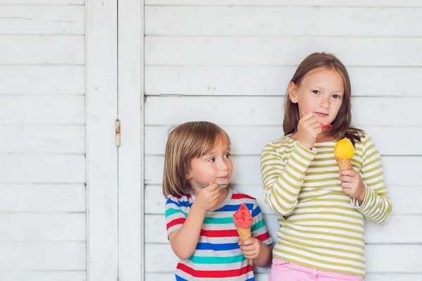 Ritratto di due adorabili bambini che mangiano gelato colorato all'aperto, in piedi accanto allo sfondo di legno bianco — Foto Stock