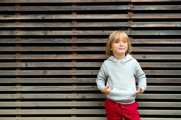 Fashion portrait of adorable toddler boy wearing grey sweatshirt and red trainings, standing against wooden background — Stock Photo, Image