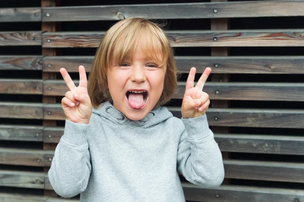 Close up portrait of a funny little boy, wearing grey sweatshirt, pulling a tongue and gesturing peace sign — 图库照片