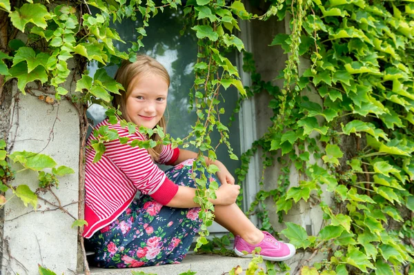 Retrato al aire libre de una linda joven sentada en el alféizar de la ventana con hiedra, con falda colorida, cárdigan de rayas rojas y blancas y zapatos sin cordones rosas —  Fotos de Stock