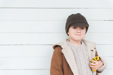 Oyuncak arabalar tutarak sıcak bej kazak, ceket ve kahverengi şapka giyen sevimli küçük bir çocuk beyaz ahşap arka plan karşı portresi