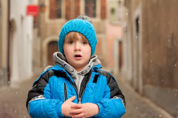 Retrato ao ar livre de um menino bonito em uma cidade sob queda de neve, vestindo casaco azul quente e chapéu de malha — Fotografia de Stock