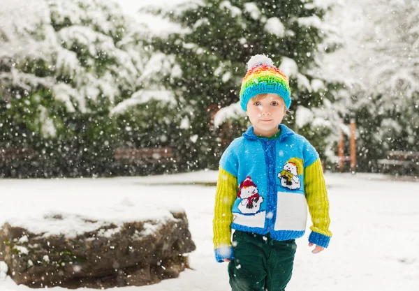 Winter portrait of a cute little boy under the snowfall, wearing christmas pullover and colorful hat — Stock Photo, Image