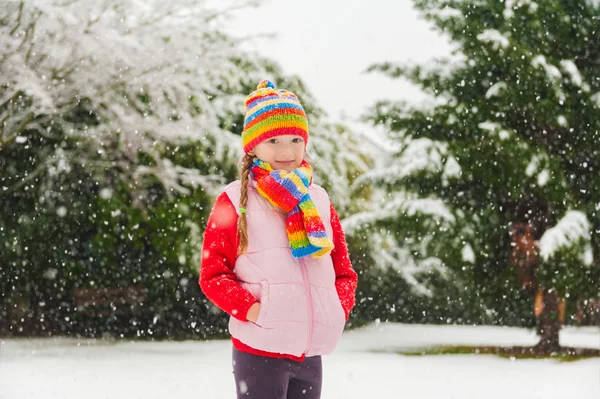 Winter portrait of a cute little girl under the snowfall, wearing red pullover, colorful hat and scarf — Stock Photo, Image