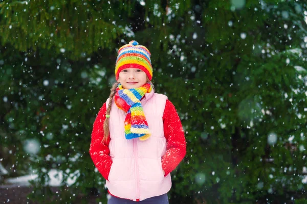 Vinter porträtt av en söt liten flicka under snöfall, klädd i röd tröja, färgglad mössa och halsduk — Stockfoto