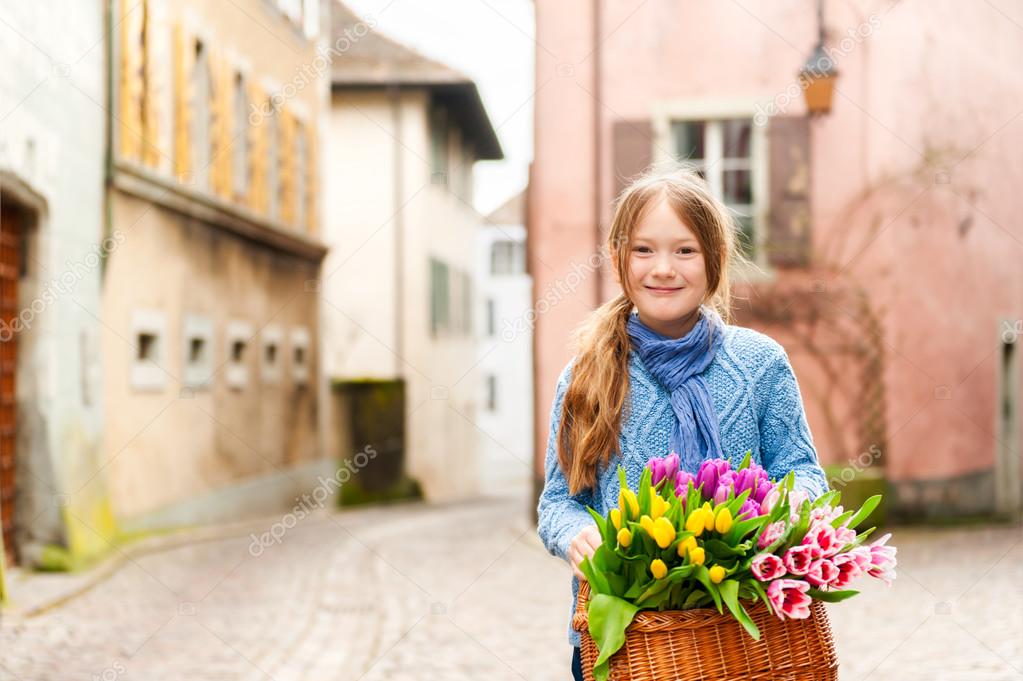 Outdoor portrait of adorable little girl of 7 years old walking in old town, holding basket full of colorful tulips, wearing warm blue pullover and scarf
